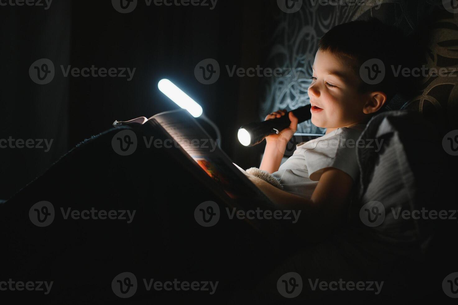 Reading book and using flashlight. Young boy in casual clothes lying down near tent at evening time photo
