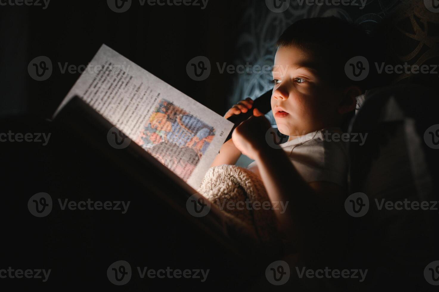 Child reading book in bed. Kids read at night. Little boy with fairy tale books in bedroom . Education for young children. Bedtime story in the evening. Cute kid under blanket in dark room with lamp photo