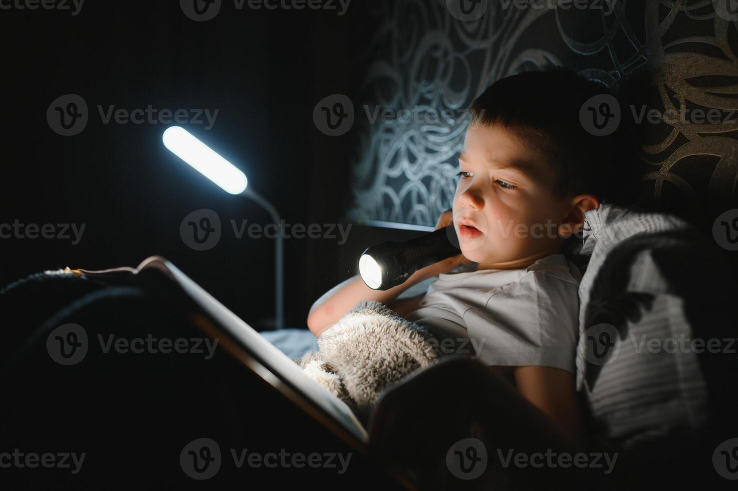 Child reading book in bed. Kids read at night. Little boy with fairy tale books in bedroom . Education for young children. Bedtime story in the evening. Cute kid under blanket in dark room with lamp photo