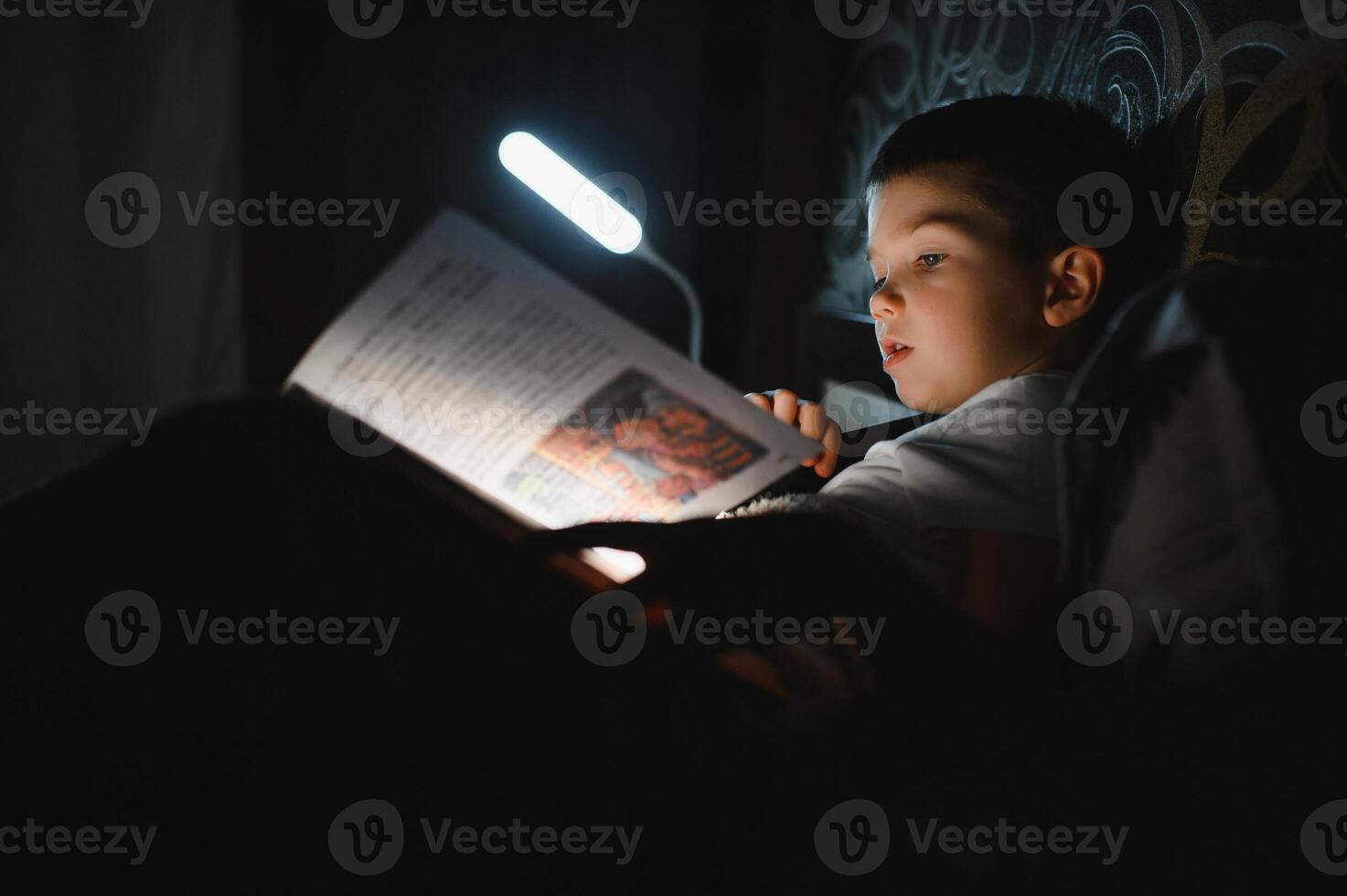 Child reading book in bed. Kids read at night. Little boy with fairy tale books in bedroom . Education for young children. Bedtime story in the evening. Cute kid under blanket in dark room with lamp photo