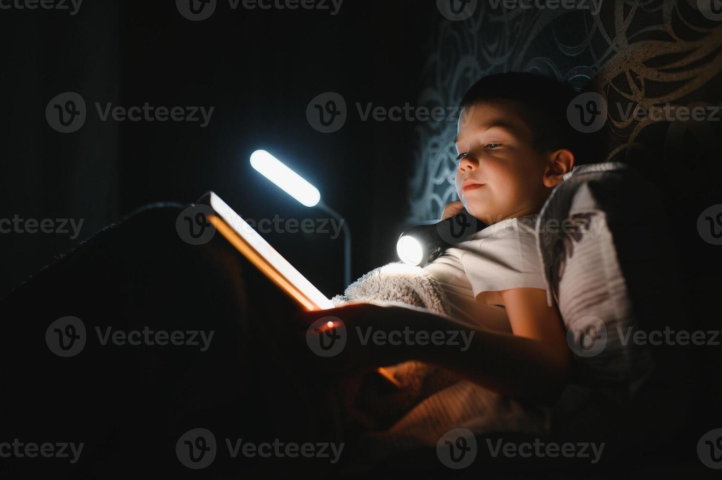 Reading book and using flashlight. Young boy in casual clothes lying down near tent at evening time. photo