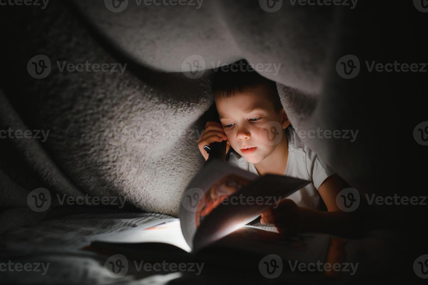 Boy with flashlight reading book under blanket at home photo