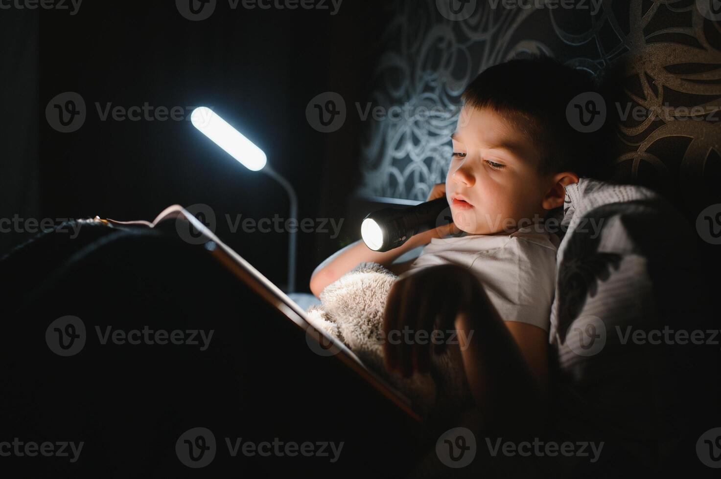 Portrait of cute little boy reading in bed with flashlight in dark room, enjoying fairytales photo