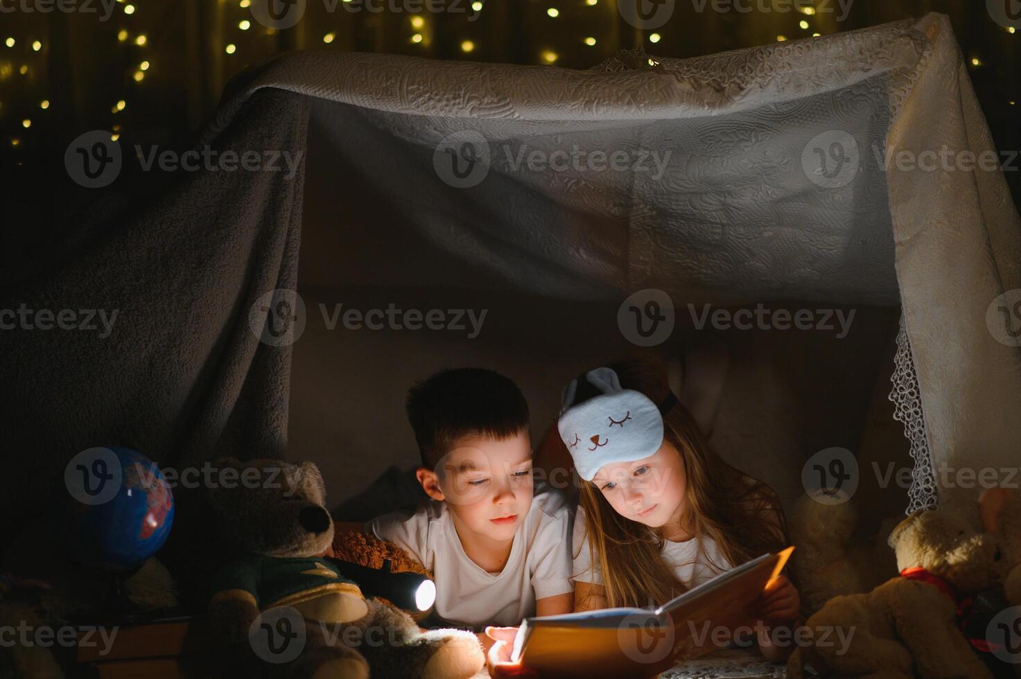 Two children with flashlight read a book under a blanket as a tent photo