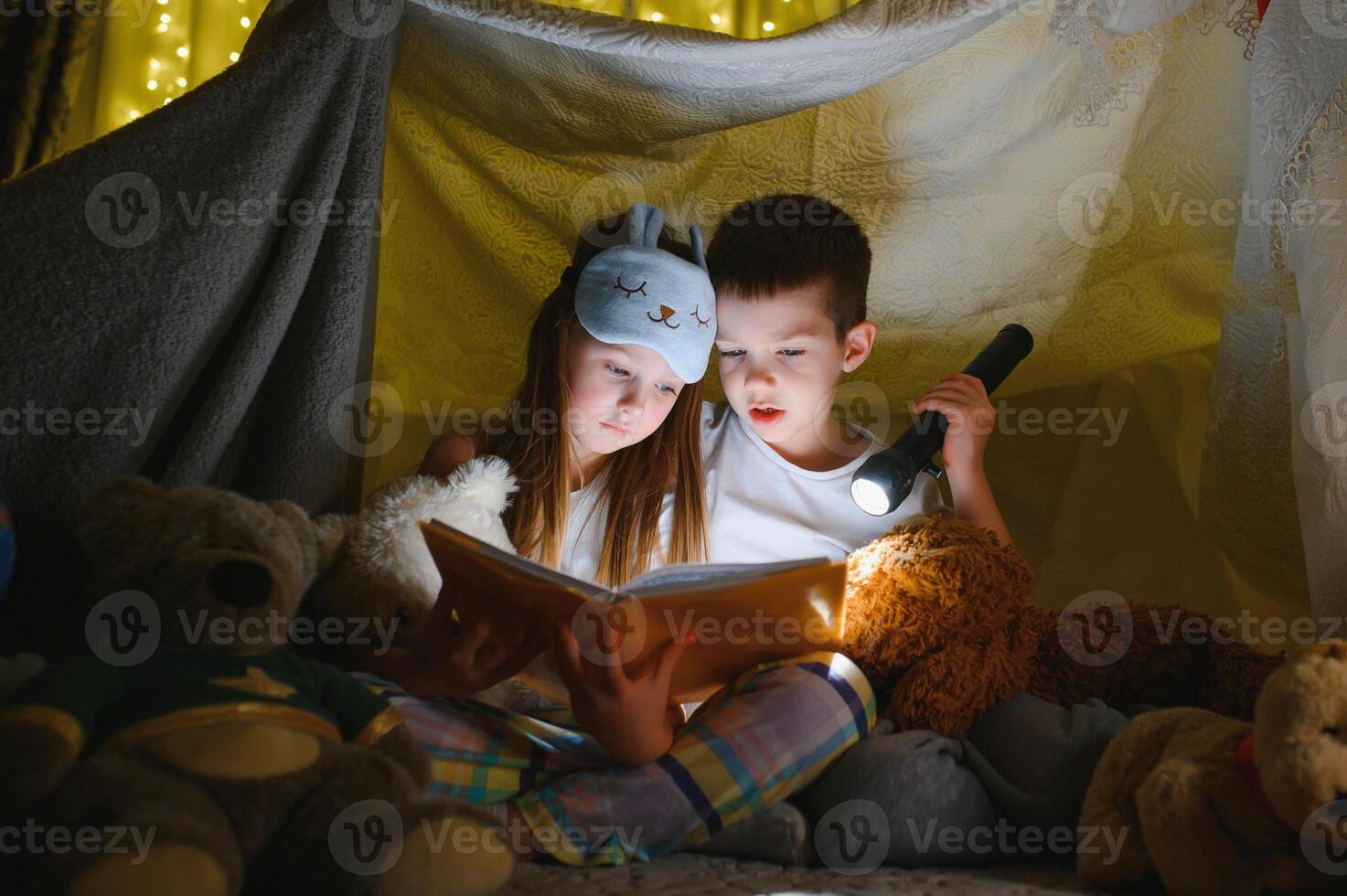 Two children with flashlight read a book under a blanket as a tent photo