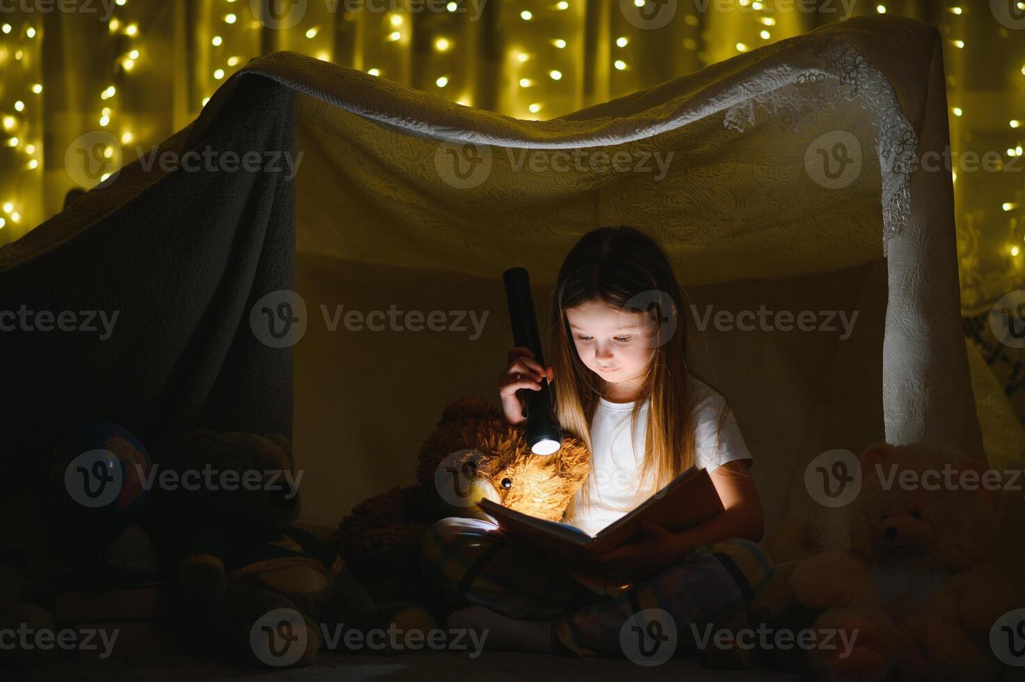 child girl reading with book and flashlight and teddy bear in tent. before going to bed photo