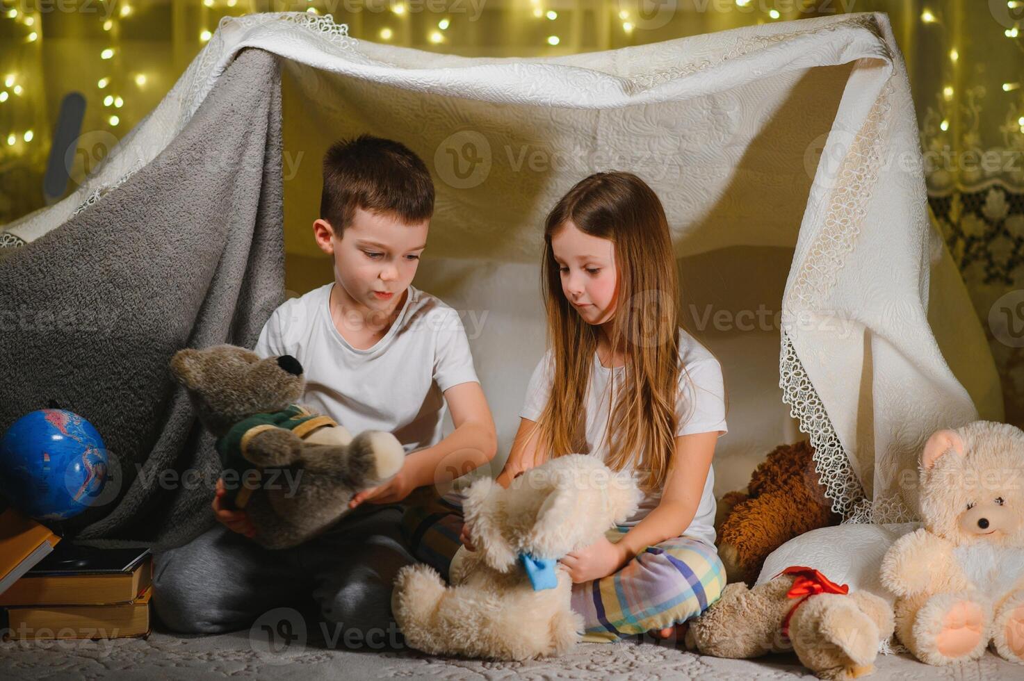 Siblings sit in a hut of chairs and blankets. Brother and sister reading book with flashlight at home photo