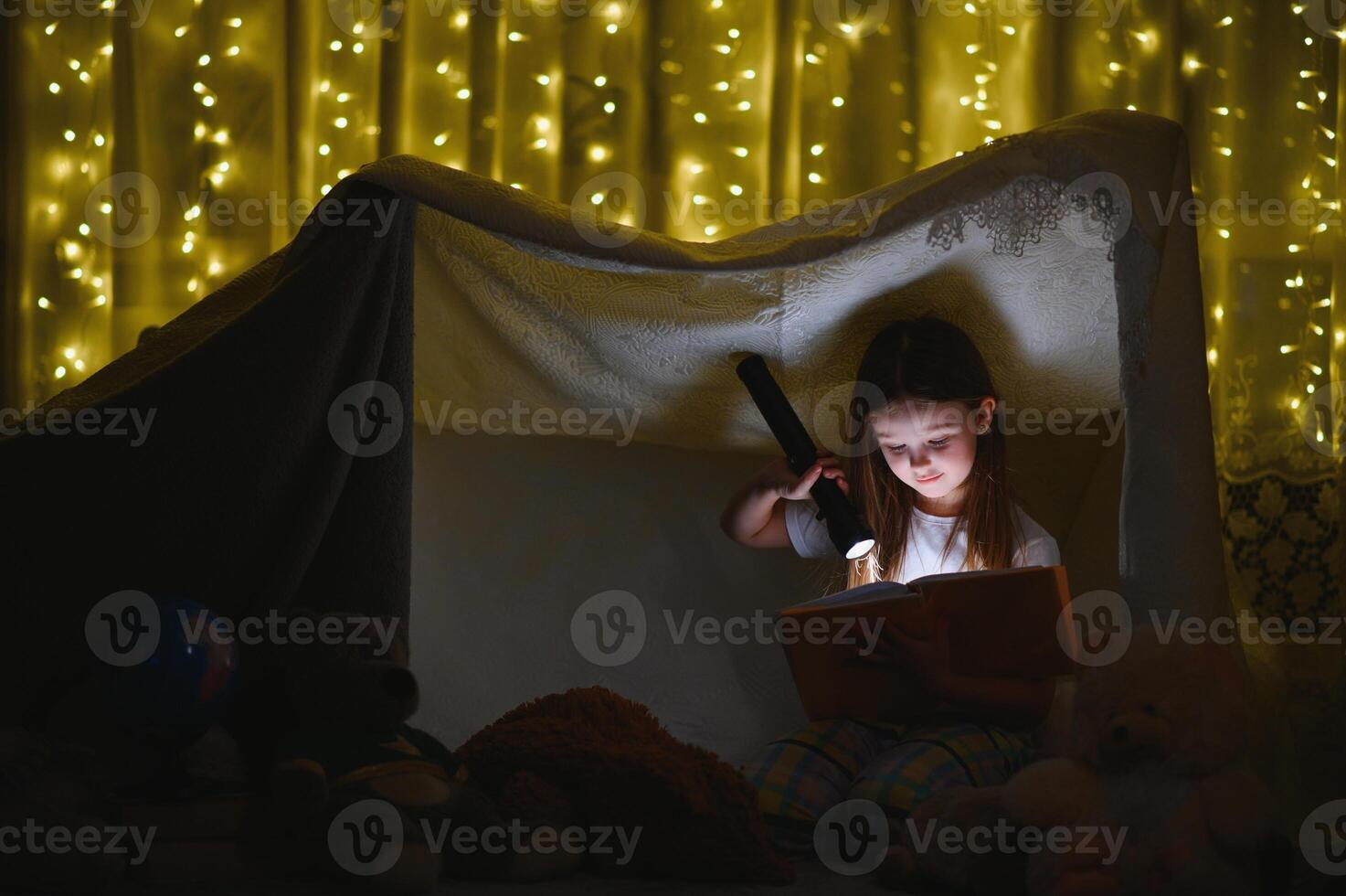 Little girl are reading a book with flashlights in tent. Happy girl playing at home. Funny lovely kid having fun in room. photo