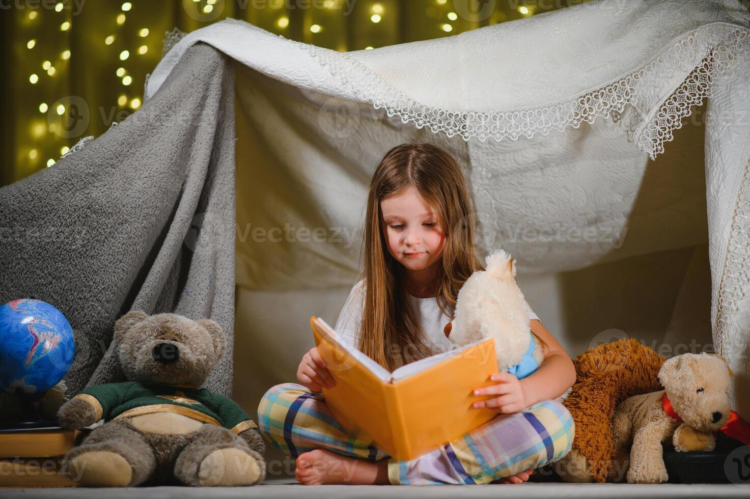 child girl reading with book and flashlight and teddy bear in tent. before going to bed photo