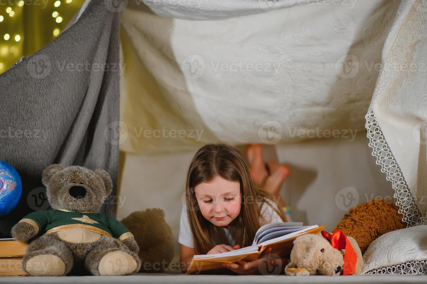 child girl reading with book and flashlight and teddy bear in tent. before going to bed photo