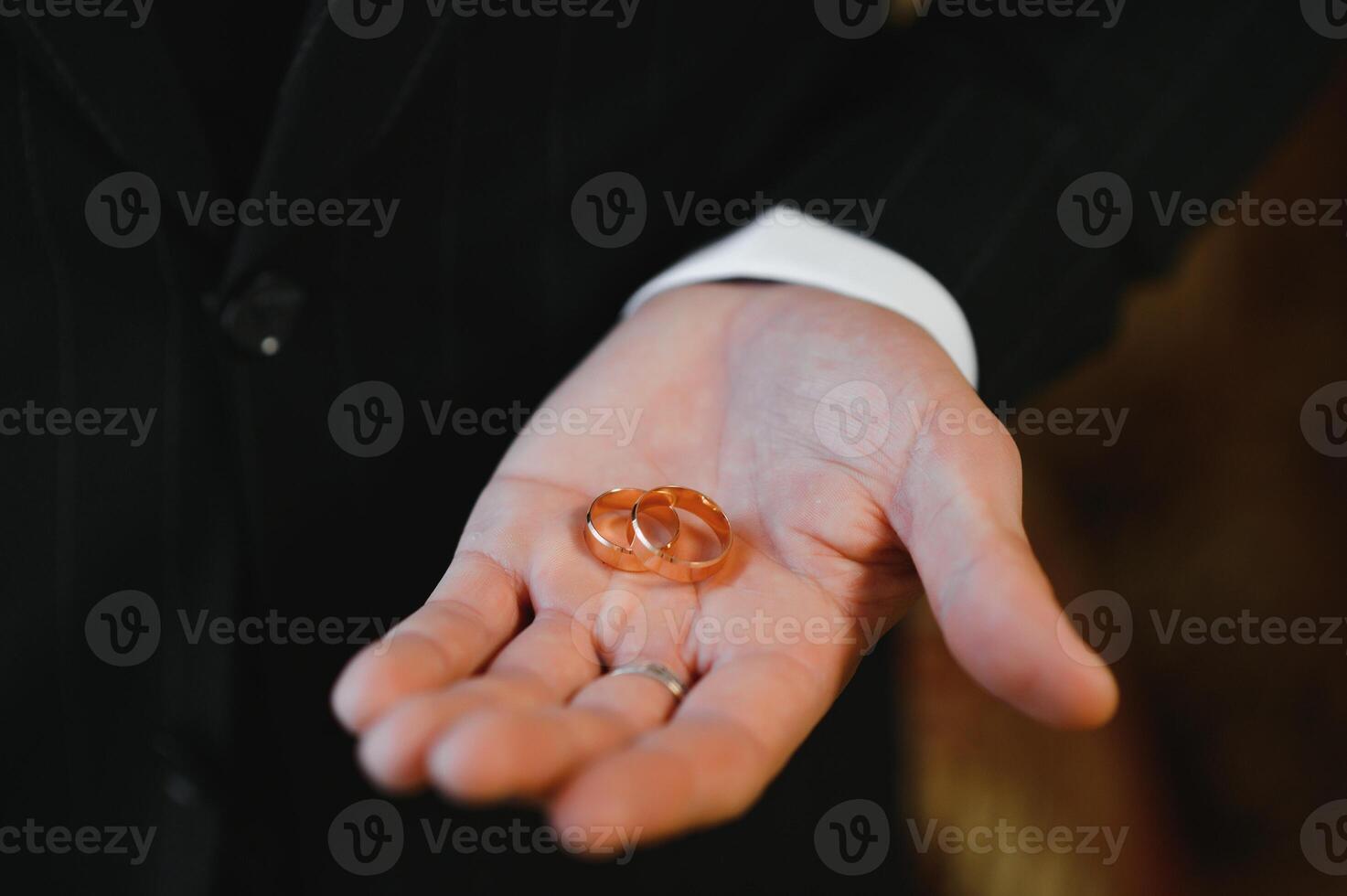 The groom on his wedding day holds two wedding rings in his hand in a hotel room. The man is wearing a white shirt and blue trousers with a vest. Male hand and wedding rings close up. photo