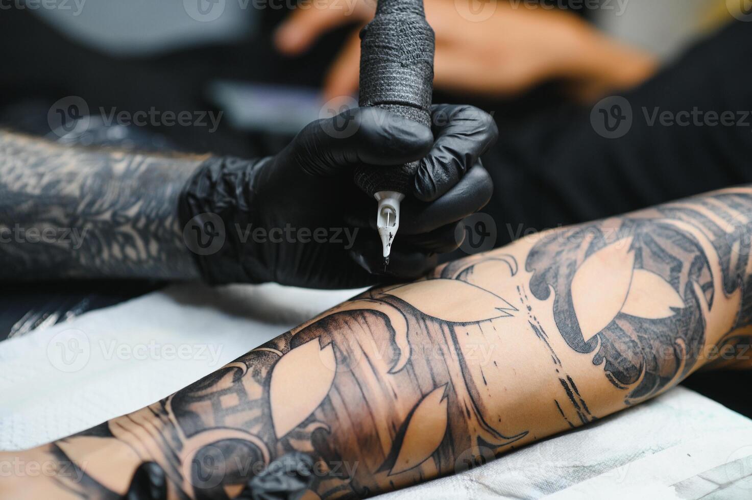Cropped close up of a bearded tattoo artist working at his studio tattooing sleeve on the arm of his male client. Man getting tattooed by professional tattooist photo
