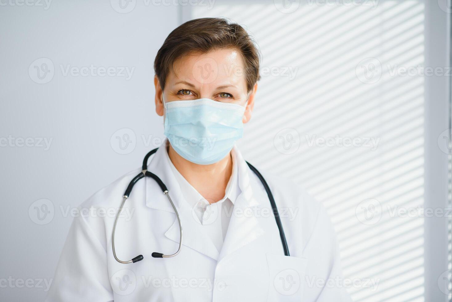 más viejo hembra médico vistiendo cara máscara y blanco médico Saco en pie en hospital. retrato foto