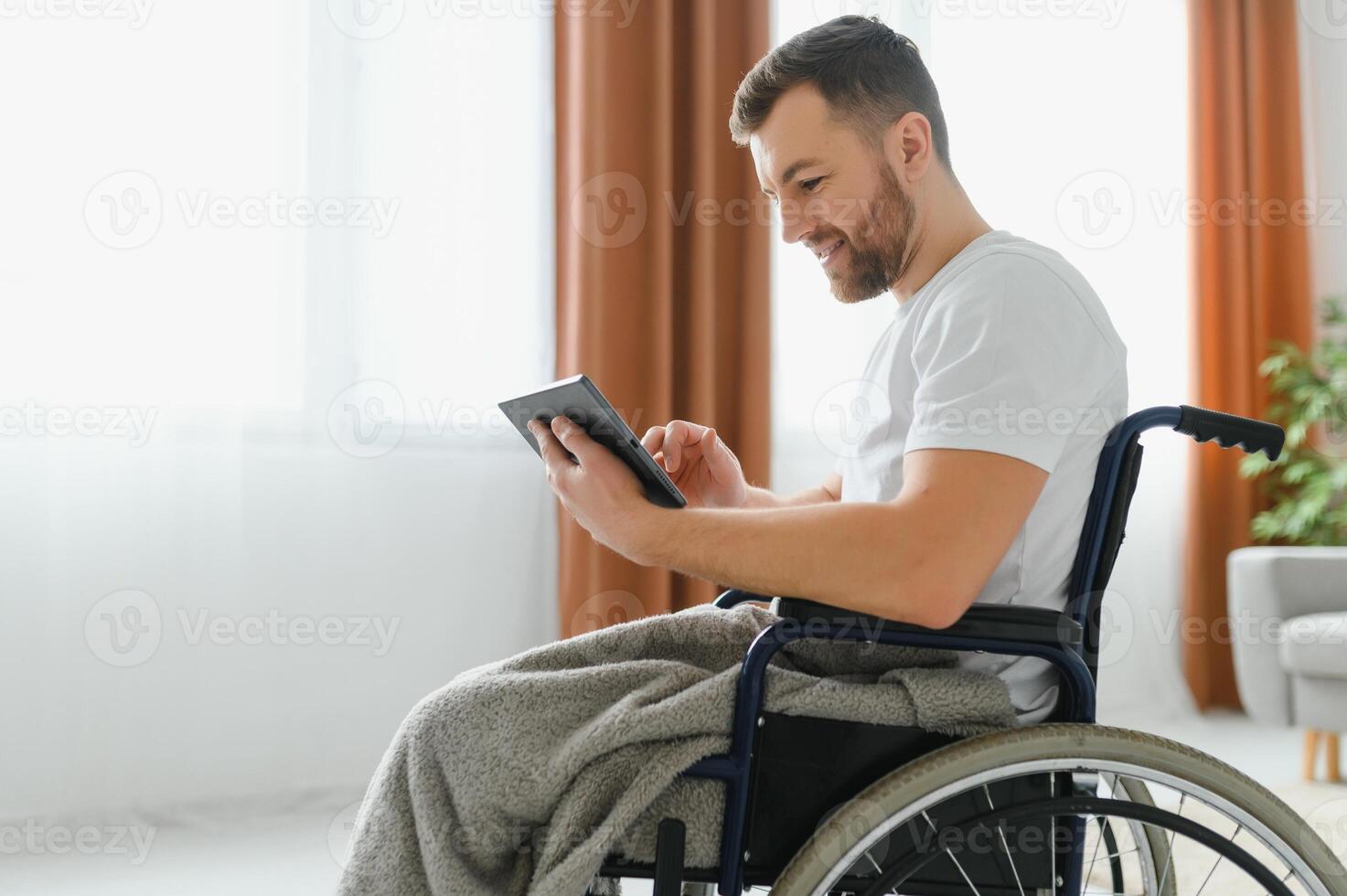 Man in wheelchair using tablet computer at home. Positive retired male with physical disability browsing web on touch pad, watching online indoors. photo