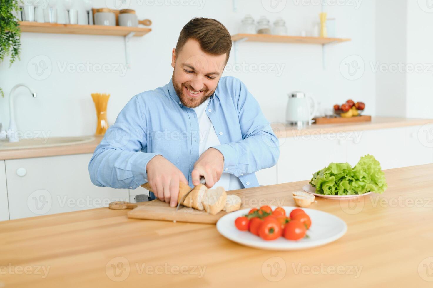 Young man sit at table and cut cucumber on salad. photo