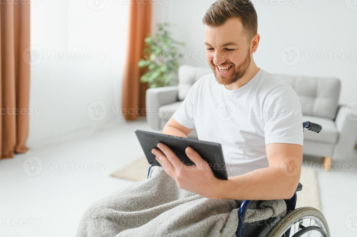 Man in wheelchair using tablet computer at home. Positive retired male with physical disability browsing web on touch pad, watching online indoors photo