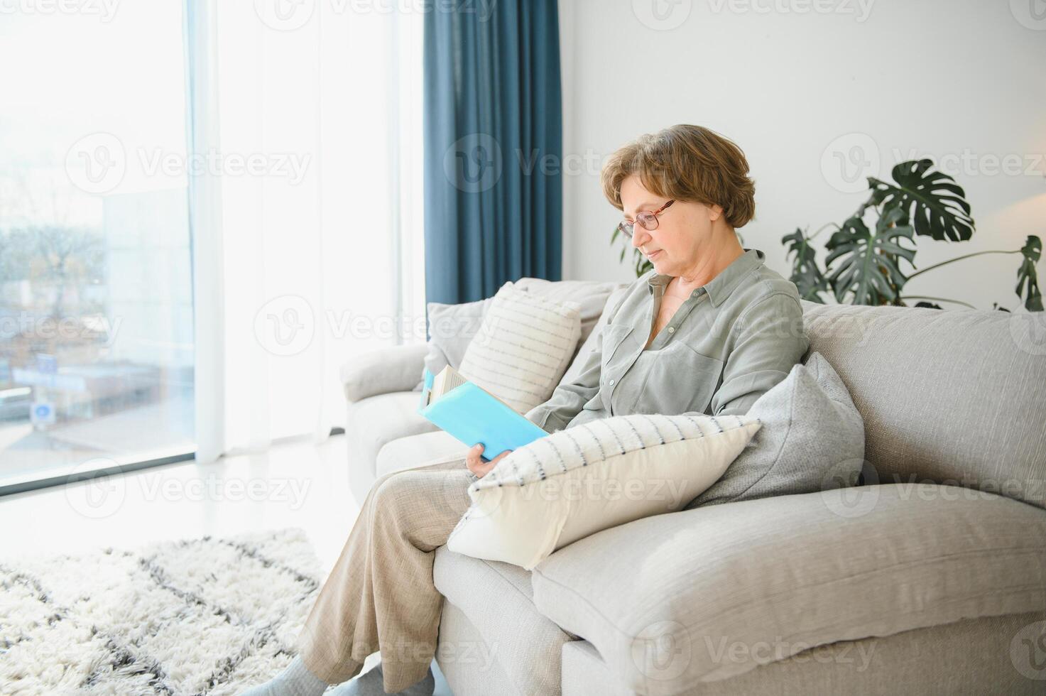 Restful reading. Portrait of thoughtful aged woman reading favorite literature at cozy home. She is lying on pillows on comfortable sofa. Legs are covered with soft blanket. photo