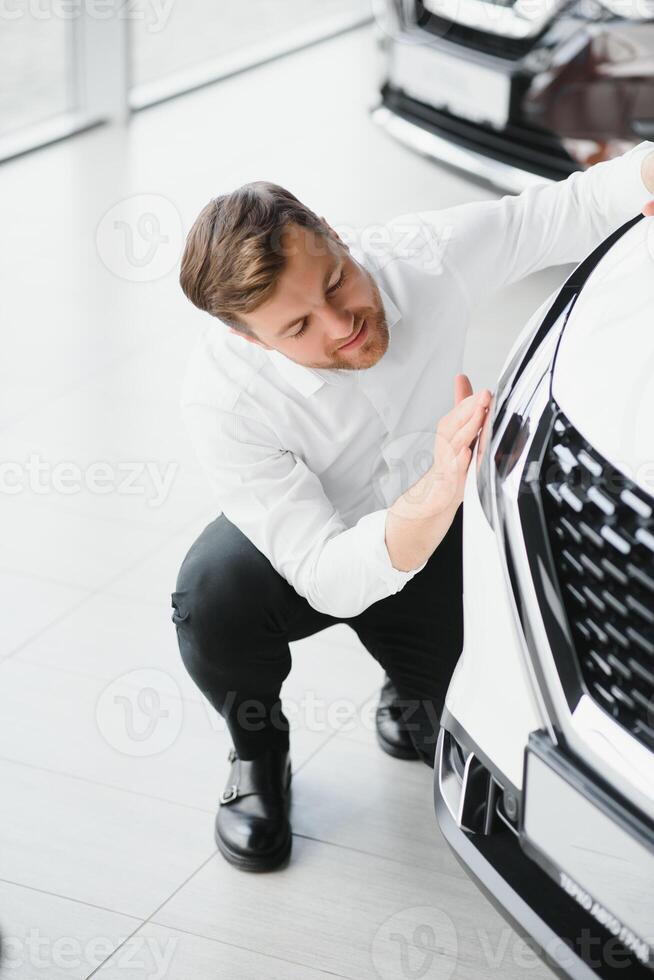 Happy handsome bearded man buying a car in dealership, guy hugging hood of new car photo