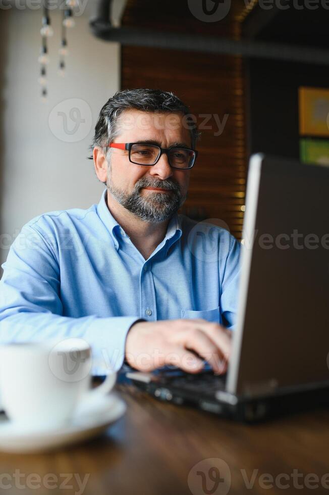 Male freelancer is working in a cafe on a new business project. Sits at a large window at the table. Looks at a laptop screen with a cup of coffee photo