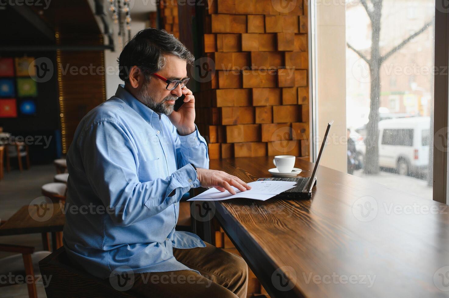 Male freelancer is working in a cafe on a new business project. Sits at a large window at the table. Looks at a laptop screen with a cup of coffee photo