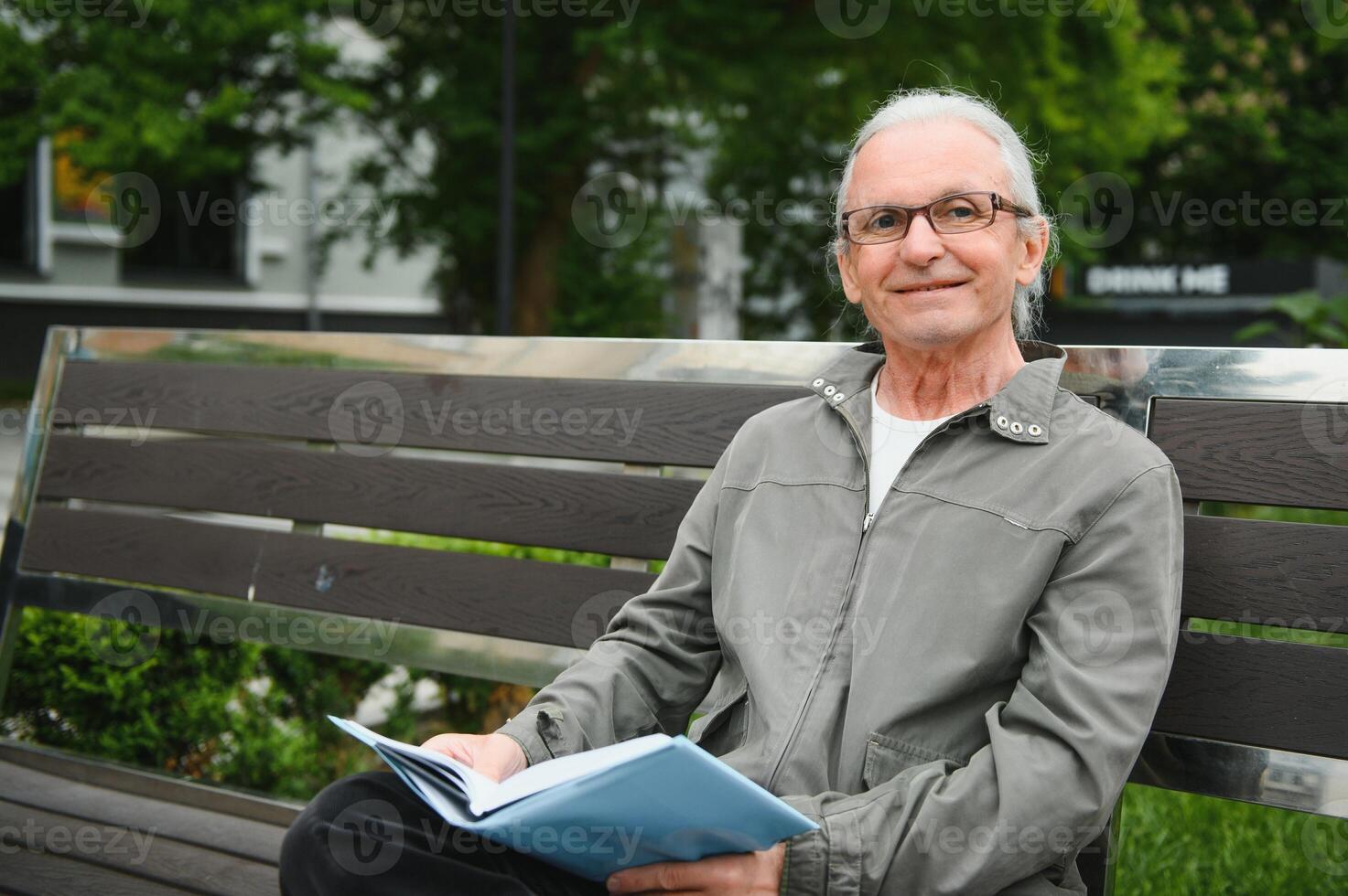 Old man with gray hair reads a book on a bench in the park. Rest in the park. photo