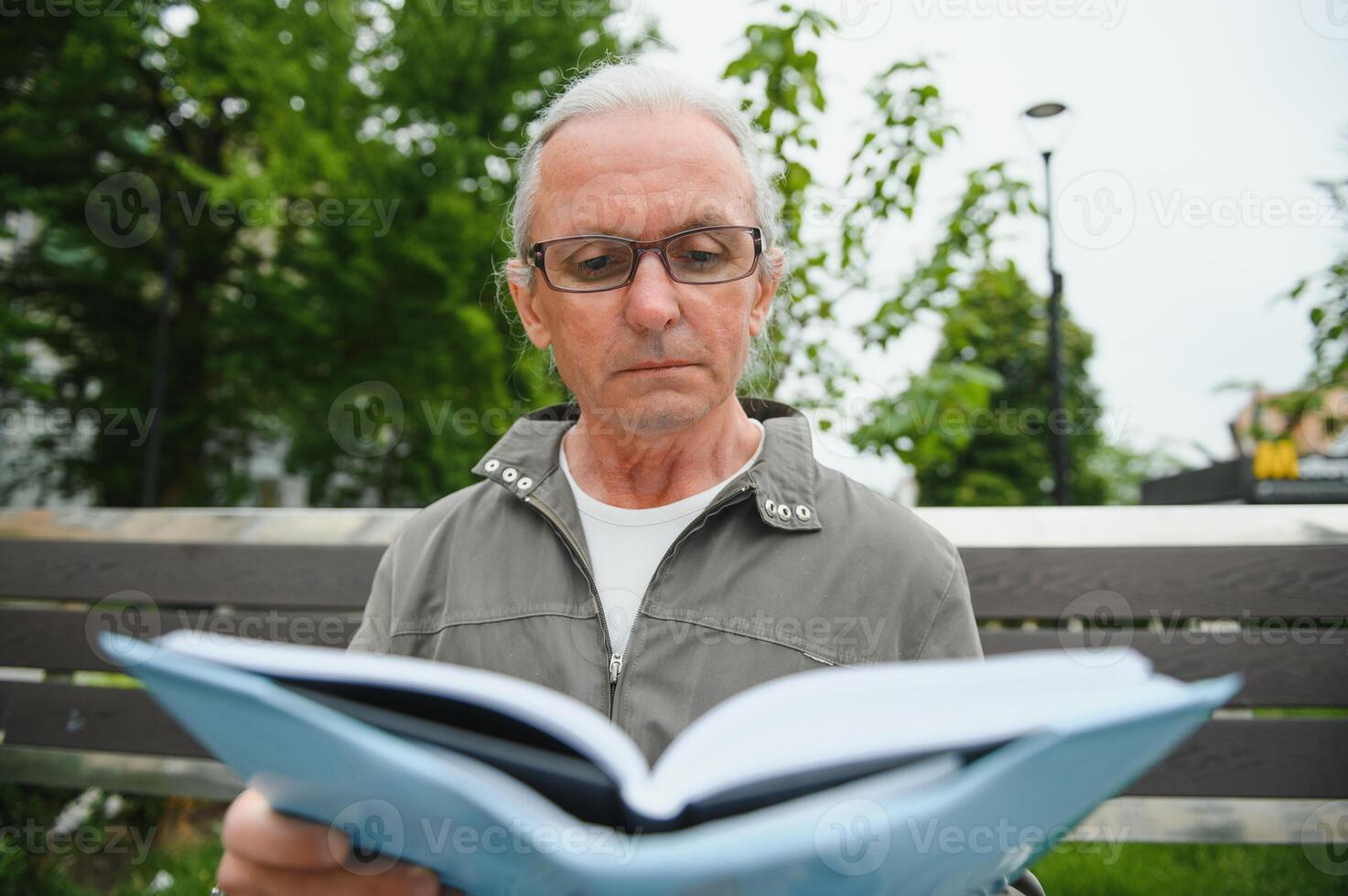 Old man with gray hair reads a book on a bench in the park. Rest in the park. photo