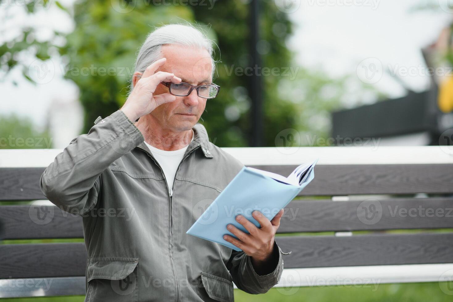 Old man with gray hair reads a book on a bench in the park. Rest in the park. photo