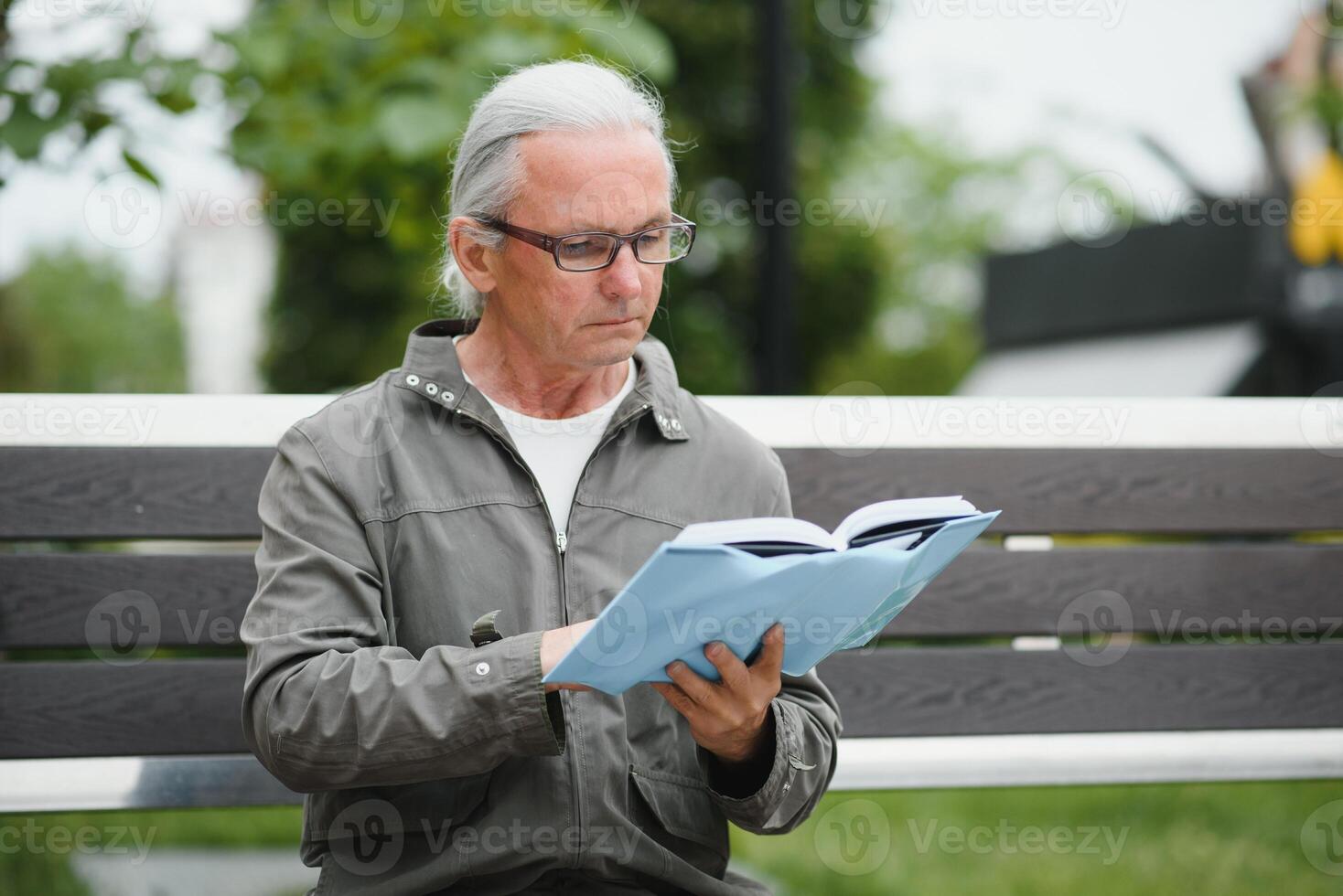 Old man with gray hair reads a book on a bench in the park. Rest in the park. photo