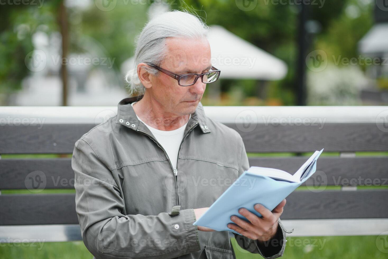 Old man with gray hair reads a book on a bench in the park. Rest in the park. photo