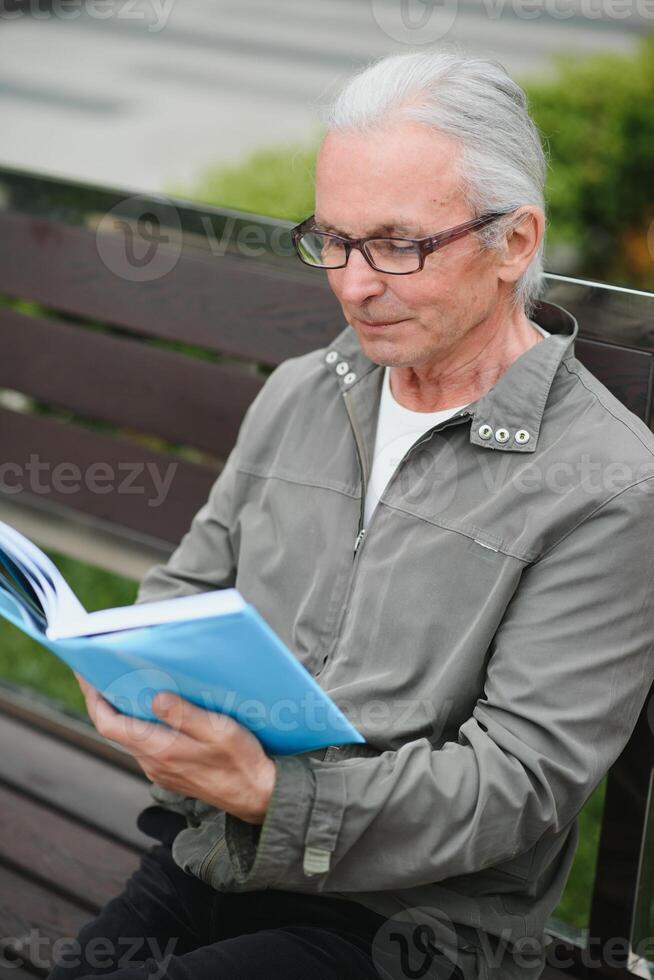 Old man with gray hair reads a book on a bench in the park. Rest in the park. photo