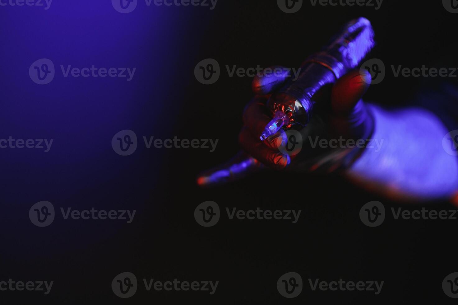 Closeup of tattooer master's hand in black glove holding machine for making tattoo art on body isolated on dark background in neon. photo