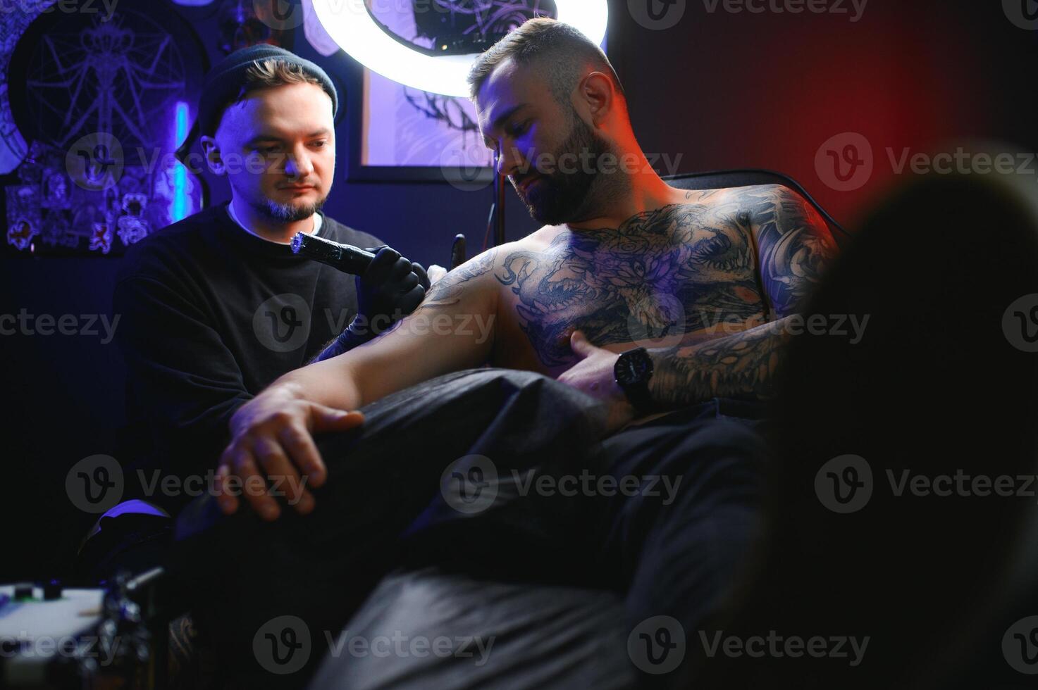bearded tattoo artist working at his studio tattooing sleeve on the arm of his male client. Man getting tattooed by professional tattooist photo