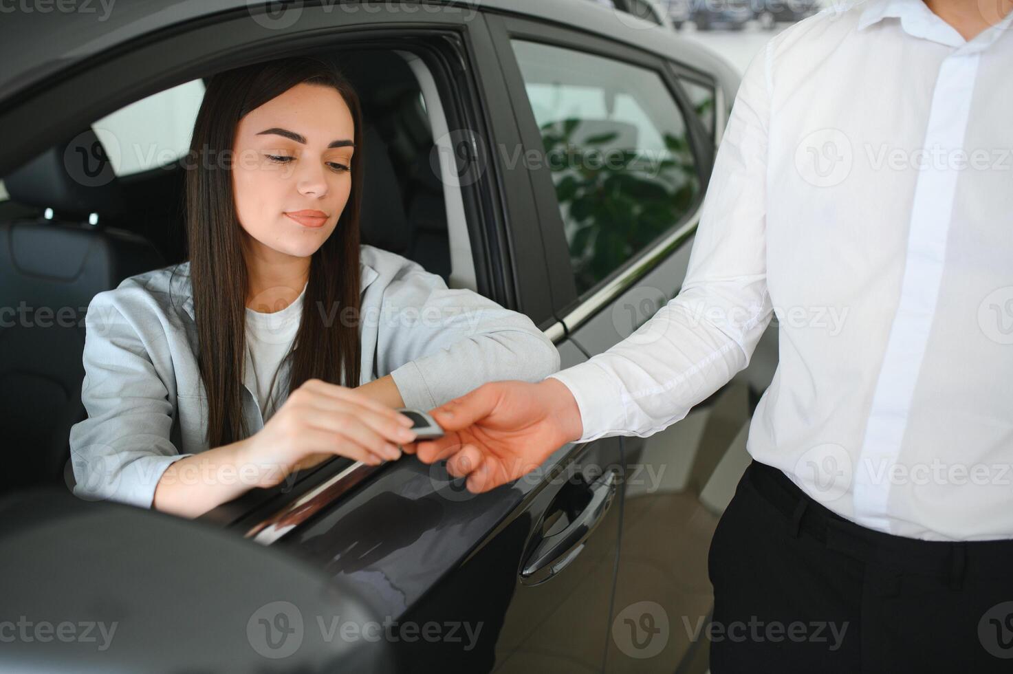 Dealer giving key to cheerful female driver while selling modern red vehicle in dealership photo