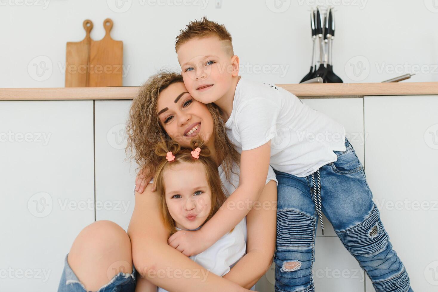 Happy mother hugging daughter and son sitting on wooden floor in modern kitchen at home. Mom hugging children. Happy family relationship concept photo