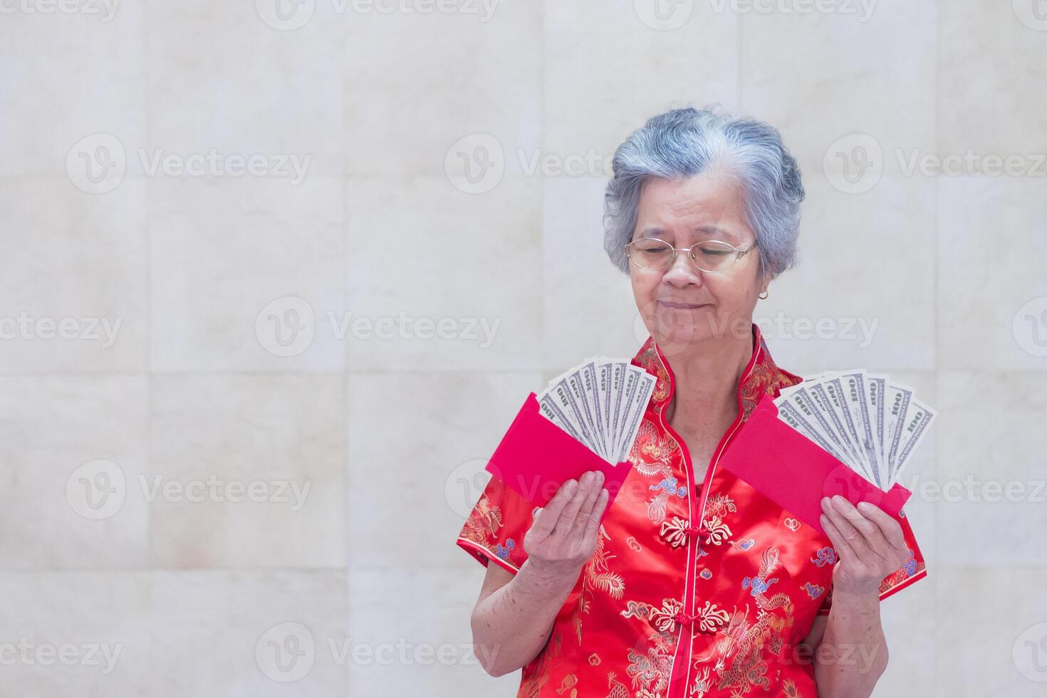 Senior woman wearing a traditional Cheongsam qipao dress holding US banknotes while standing in the living room. Concept of aged people and Chinese new year photo