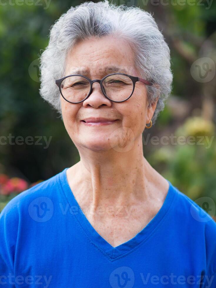 A portrait of a senior woman with short gray hair, wearing glasses, smiling, and looking at the camera while standing in a garden. Space for text. Concept of aged people and healthcare photo