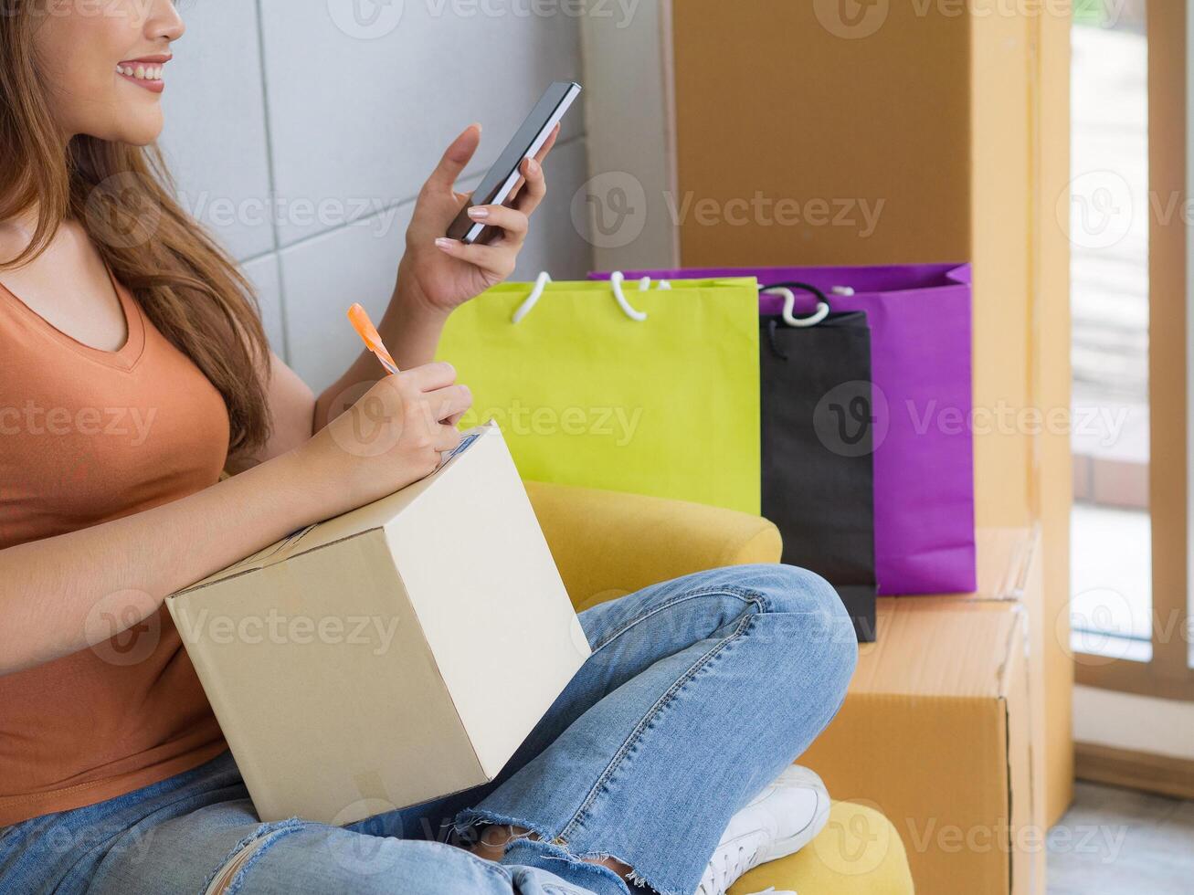 Asian businesswoman is an e-commerce entrepreneur using a smartphone and writes customer address on a parcel box while sitting on a sofa in the office. Concept of e-commerce photo