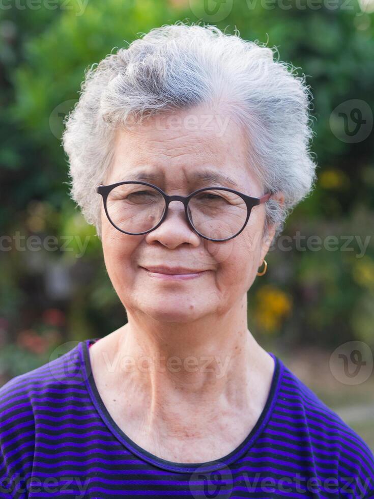 Portrait of an elderly Asian woman with short gray looking to the camera while standing in a garden. Space for text. Concept of aged people and healthcare photo