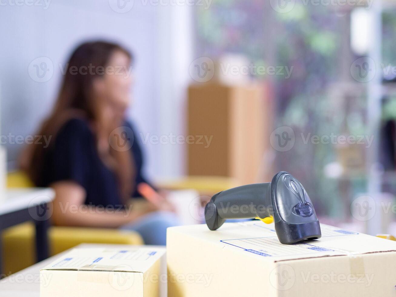 Close-up of parcel boxes and a barcode reader on the table with a young woman working while sitting on a sofa in the office a blurred background. Concept of business and e-commerce photo
