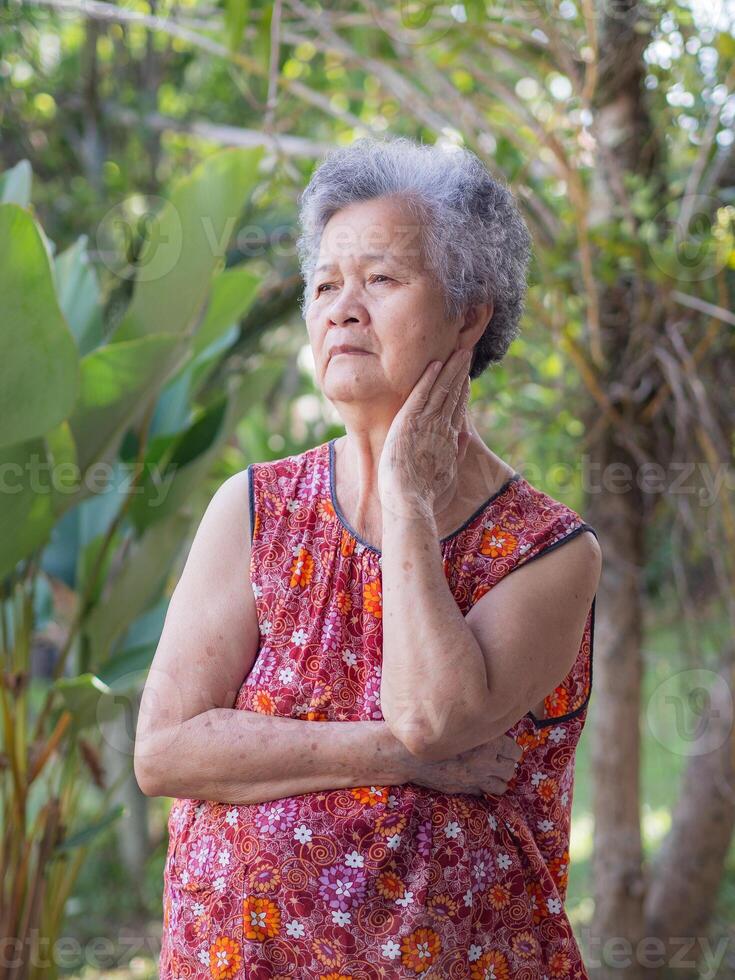 Senior woman with short gray hair hand touch the face and looking up while standing in a garden. Space for text. Concept of aged people and healthcare photo