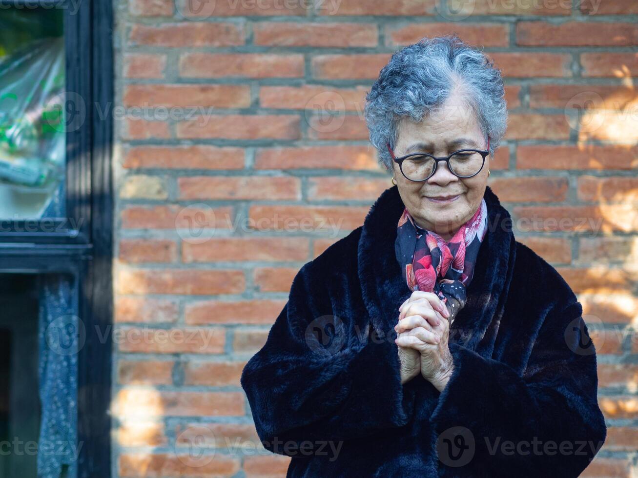 Portrait of an elderly Asian woman standing with eyes closed and praying with a brick wall background. Space for text. Concept of aged people and religion photo