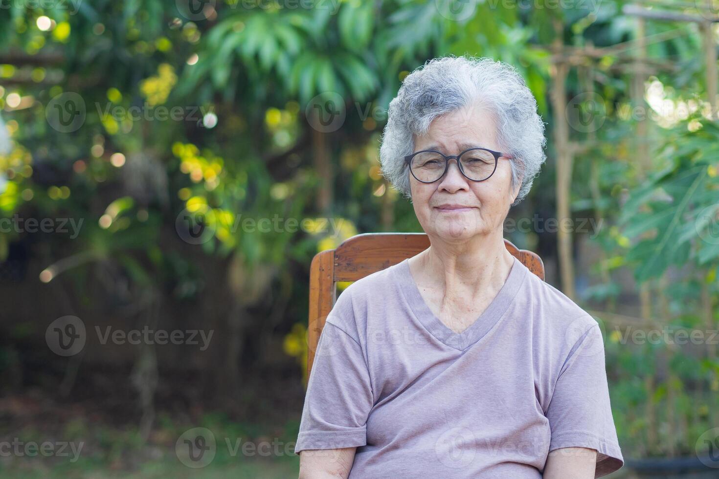 An elderly woman with short gray hair smiling and looking at the camera while sitting on a chair in the garden. Space for text. Concept of aged people and healthcare photo