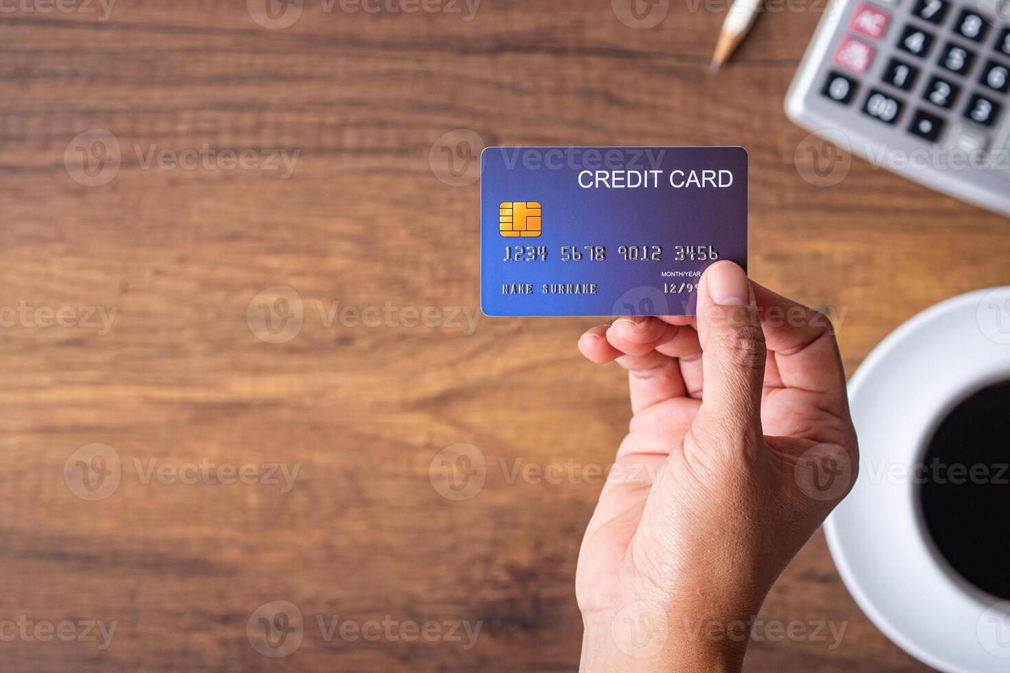 Close-up of hand holding a blue credit card with a white coffee cup, pencil, and calculator placed on wooden table. Selective focus. Space for text. Business and payment concept photo