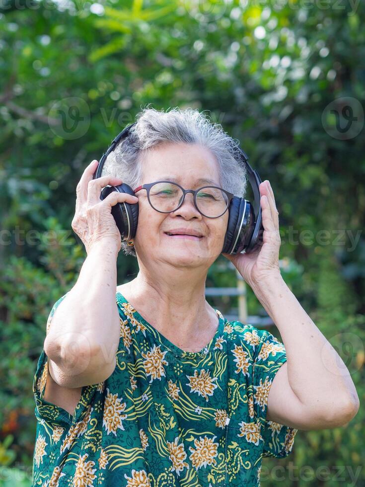 Senior woman with short gray hair wearing wireless headphones listening to a favorite song, smiling while standing in a garden. Concept of aged people and realxation photo