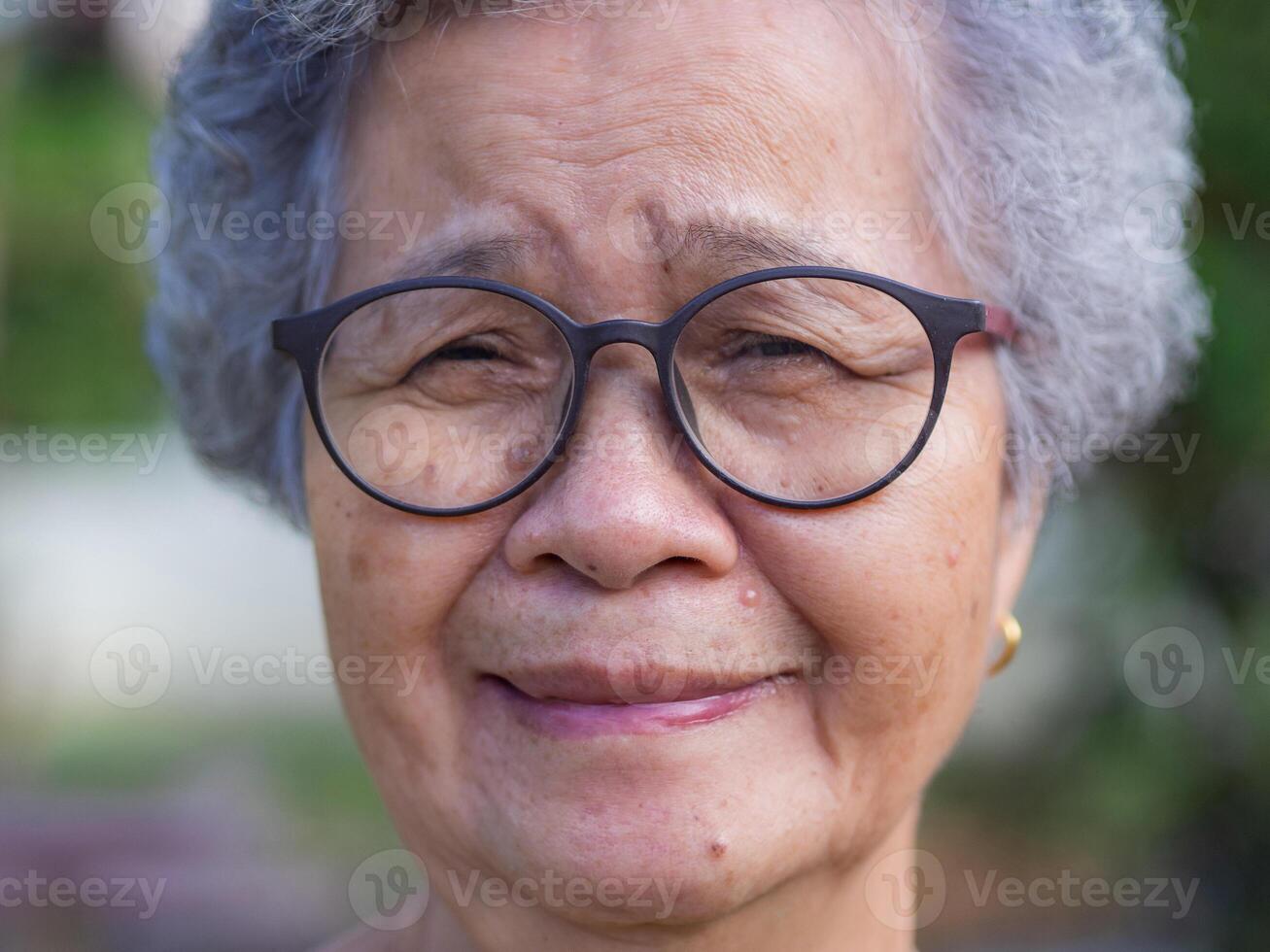 Close-up of face senior woman with short gray hair, wearing glasses, smiling and looking at the camera while standing in a garden. Concept of aged people and healthcare photo