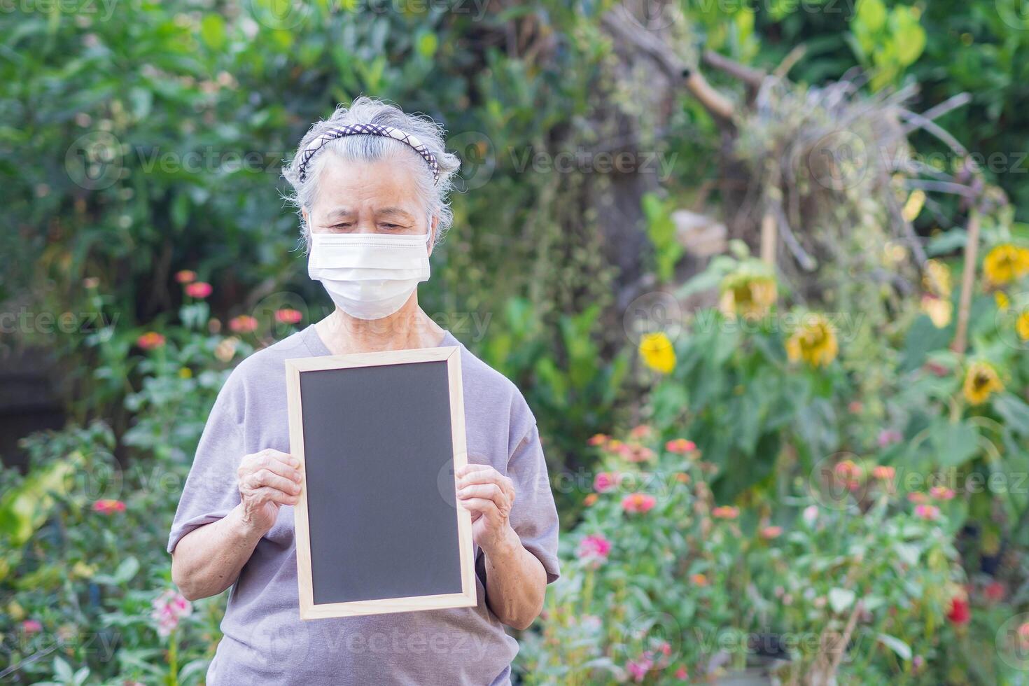 Portrait of an elderly woman wearing a face mask and holding a blackboard while standing in a garden. Space for text. Concept of aged people and healthcare photo