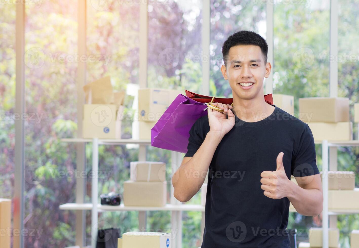 Cheerful young man holding colorful shopping bags, giving thumb up, and looking at the camera while standing in the office with sunlight. Concept of business and e-commerce photo