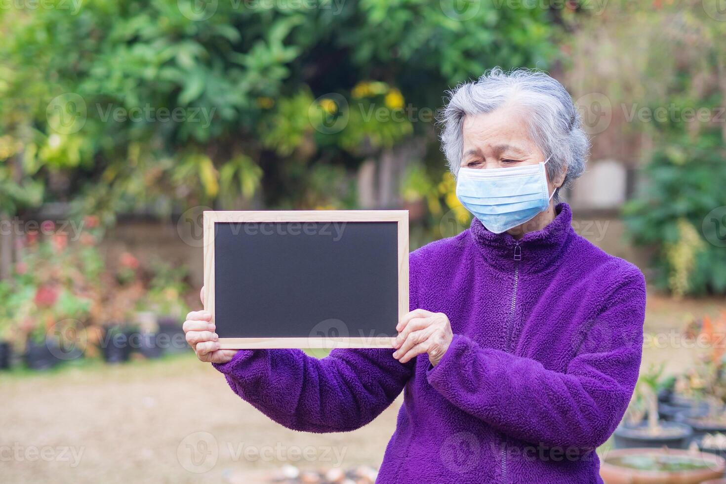 A senior woman wearing a face mask and holding a blackboard while standing in a garden. Mask for protection virus, covid-19, coronavirus, bacteria, dust. Concept of aged people and healthcare photo