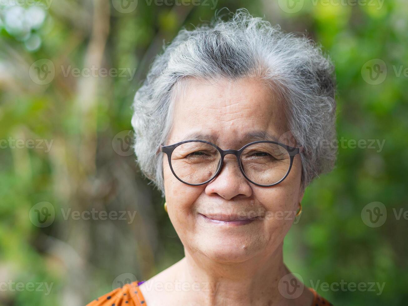 Portrait of a senior woman with short gray hair, wearing glasses, smiling, and looking at the camera while standing in a garden. Space for text. Concept of aged people and healthcare photo