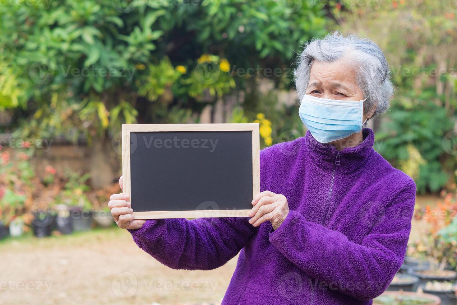 Senior woman wearing a face mask, holding a blackboard, and looking at the camera while standing in a garden. Mask for protection Covid-19, virus, bacteria, etc. Concept of healthcare photo